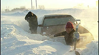 Two men with shovels attempt to dig a sedan out of a deep snow bank