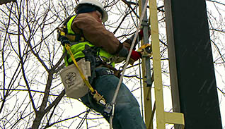 A man wearing a green safety vest climbs a ladder he is tethered to for fall prevention
