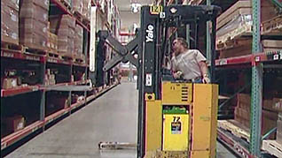 A man driving a reach truck reaches for inventory in an aisle full of boxes