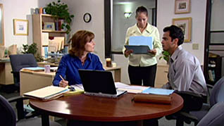 A man and a woman sit at a desk looking over papers while a woman holding folders delegates tasks to them